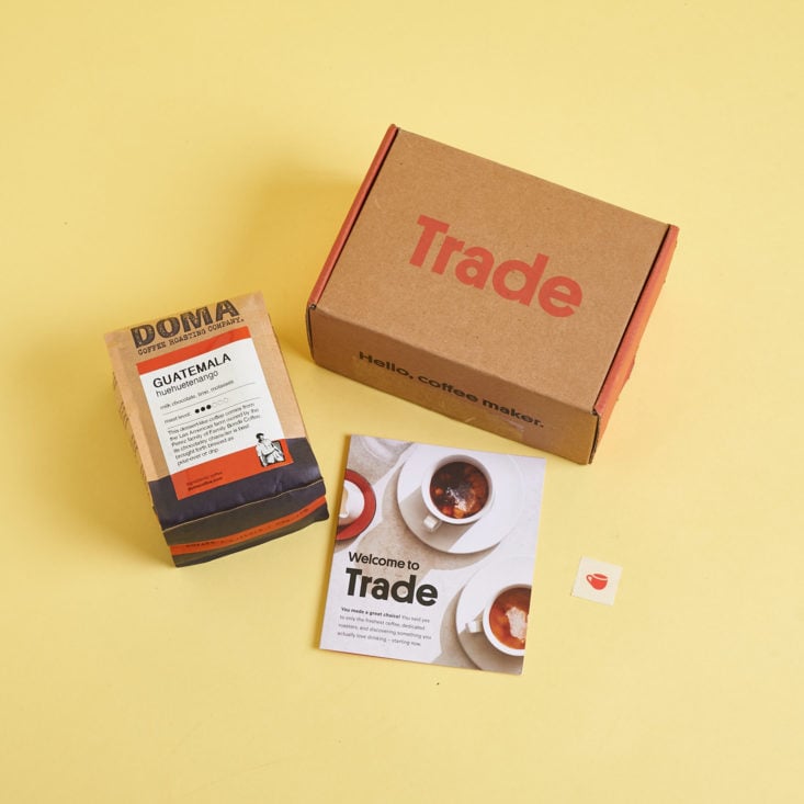 A bird's eye view of the Trade Coffee cardboard box, 1 package of coffee beans, and an informational booklet and sticker.