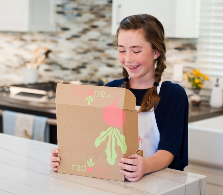 12-year-old girl with a white aprong and pigtails opening the Raddish Kids box at the counter in a kitchen.