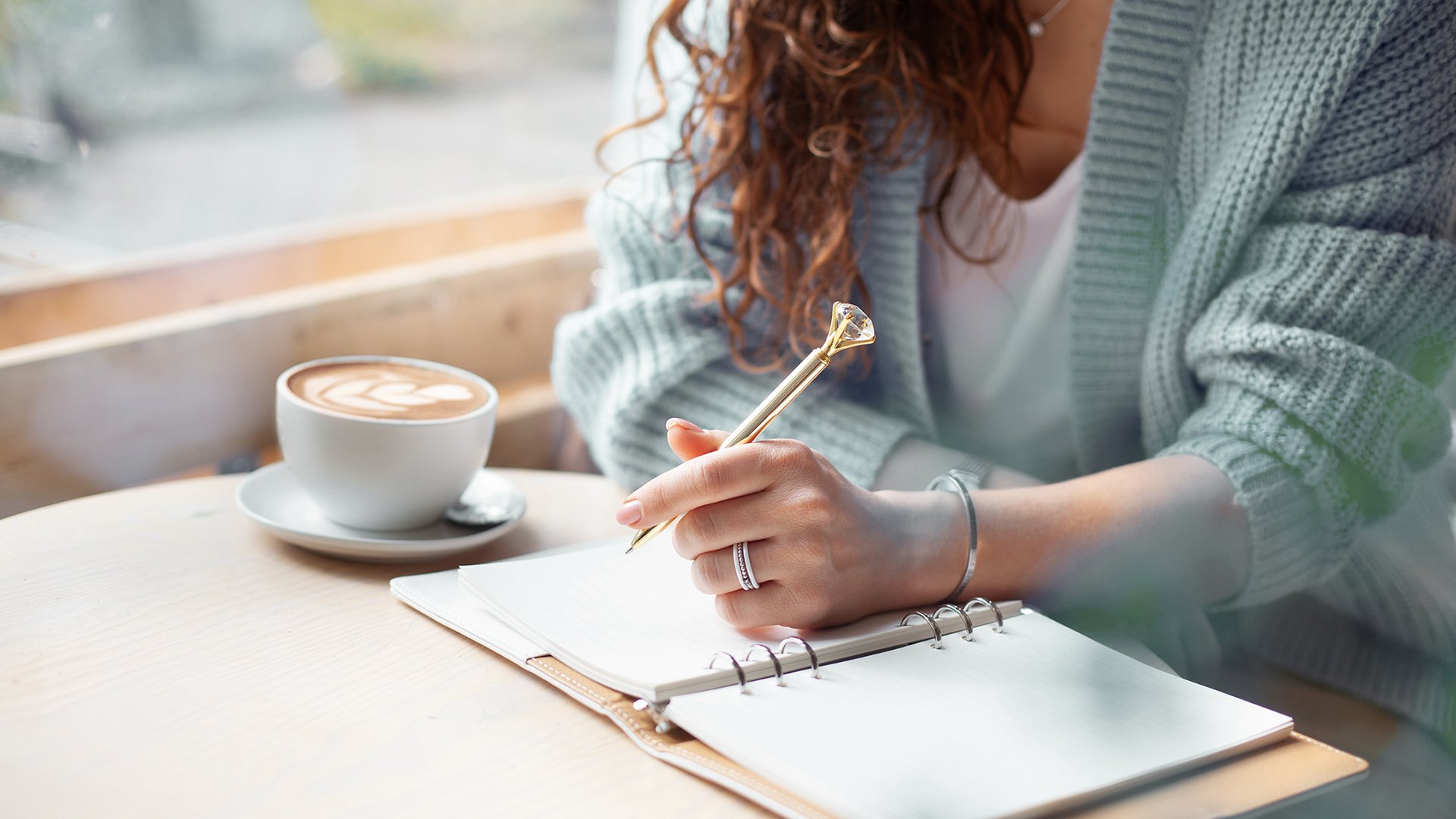 Young woman in blue warm sweater sitting near the big window of coffee shop and writing notes