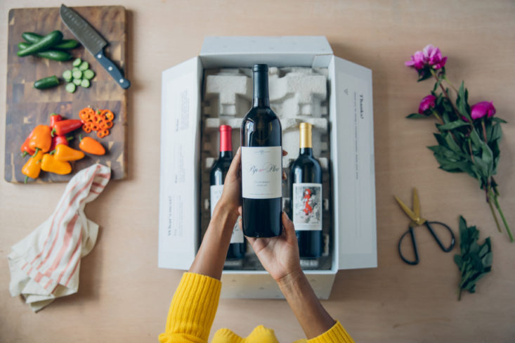 hands holding a wine bottle in the air over a box filled with wine, on the table beside it are vegetables and flowers