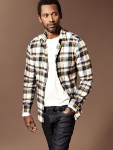 man wearing white shirt and open flannel shirt in front of a brown background