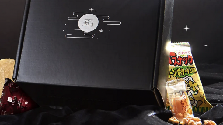 bokksu black subscription box perched on a black background with snacks surrounding it