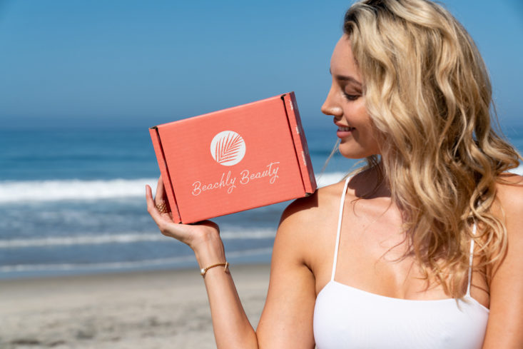 woman with blonde hair in a white tank top standing on the beach holding an orange beachly box