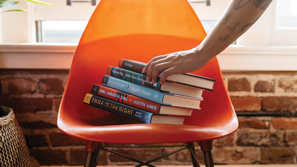 Photo of a hand choosing a book from a stack of books in an orange chair