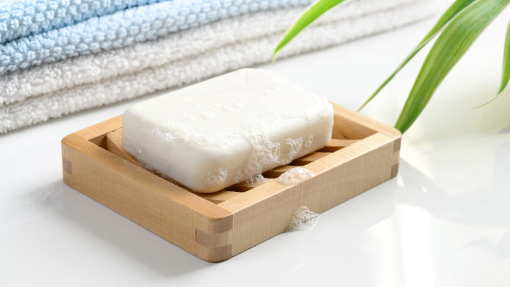 White Soap bar with foam on wooden soap dish and cotton towels on white table.