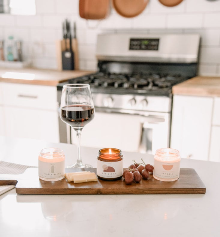 vellabox candles lined up on a charcuterie board with a glass of wine, grapes, and cheese set on a kitchen counter