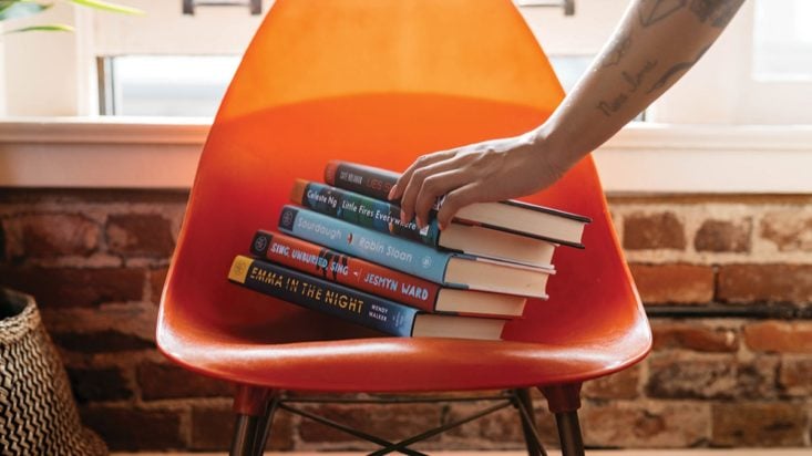 photo of orange chair filled with five books and someone picking up the top one