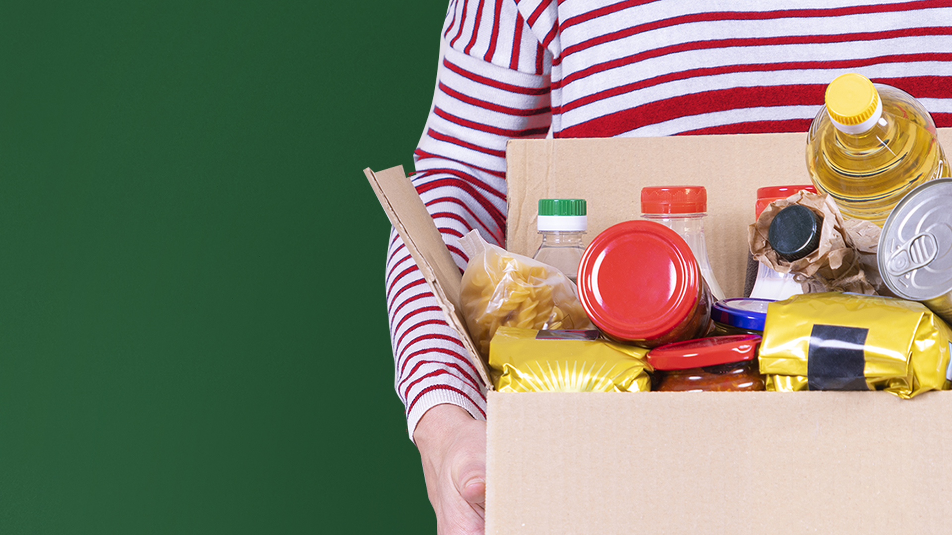 Volunteer hands holding food donations box with grocery products on white desk.