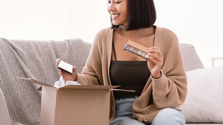 Beauty Box Concept. Portrait of smiling asian lady received delivery package, holding eyeshadow palette for makeup, unpacking cardboard parcel with cosmetics product, sitting on the couch at home