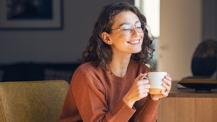 Happy young woman drinking a cup of tea in an autumn morning. Dreaming girl sitting in living room with cup of hot coffee enjoying under blanket with closed eyes. Pretty woman wearing sweater at home and enjoy a ray of sunshine on a winter afternoon.